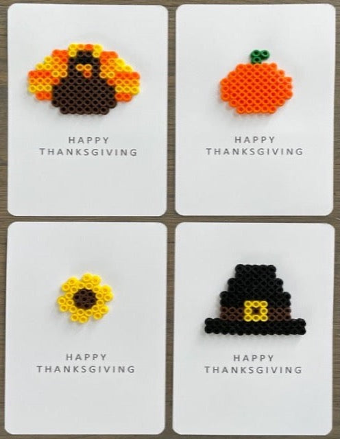 Picture of a set of 4 Happy Thanksgiving cards. One card has a dark brown, orange, and yellow turkey on it, one has an orange pumpkin on it, one has a yellow sunflower on it, and one has a black with yellow buckle pilgrim hat on it