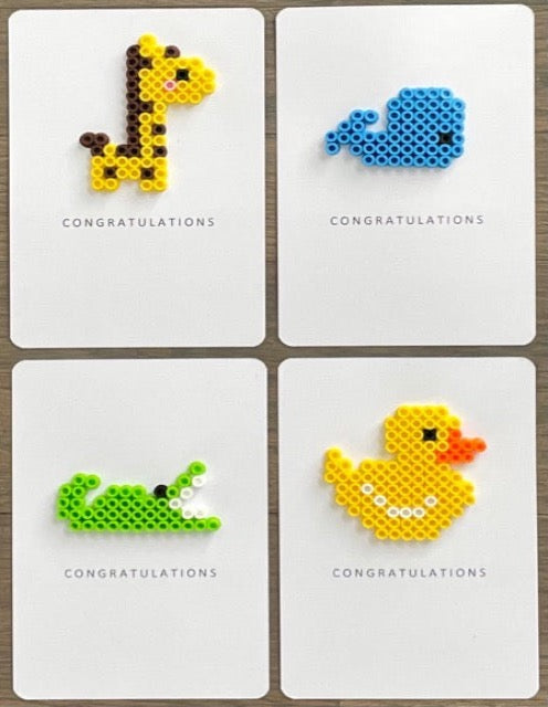 Picture of a set of 4 congratulations cards. One yellow and dark brown giraffe, one blue whale, one lime green alligator, and one yellow duck.