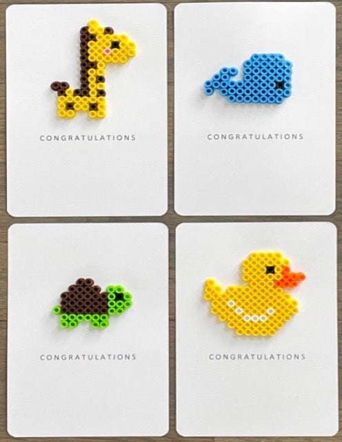 Picture of a set of 4 congratulations cards. One yellow and dark brown giraffe card, one blue whale card, one lime green and dark brown turtle card, and one yellow duck card.
