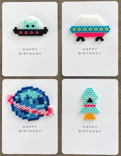 Picture of a set of 4 Space theme Happy Birthday cards. One card is black and pink spacecraft with light blue alien, one has a pink, blue, and yellow spacecraft, one has a pink and blue planet, one has a light blue and pink rocket 