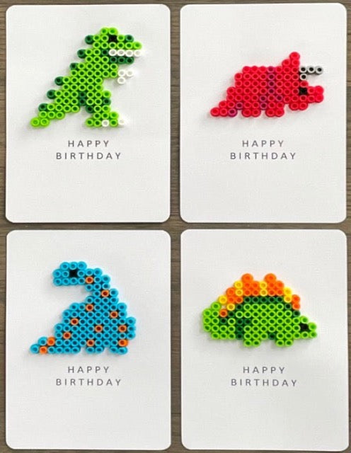 Picture of a set of 4 happy birthday cards the includes one lime green and dark green dinosaur, one red dinosaur, one blue and orange dinosaur, and one lime green, orange, and yellow dinosaur 