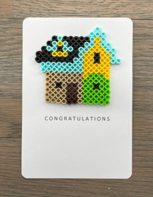 Picture of a congratulations card with a multi-colored house on it. House is tan, lime green, yellow, light blue with a black roof and dark brown door