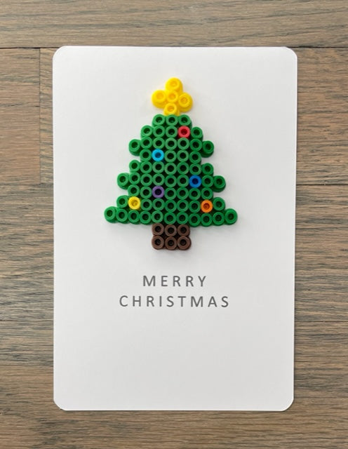 Picture of a Merry Christmas card with green Christmas tree on it that has a dark brown truck, yellow tree topper, and multi-colored ornaments
